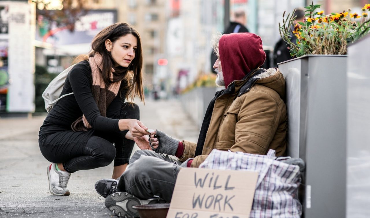 A young woman giving money to homeless beggar man sitting outdoors in city.